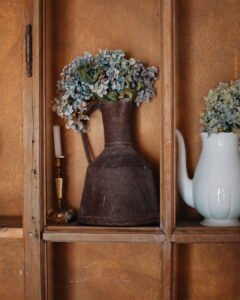 An antique dark metal pitcher filled with blue hydrangeas next to a white ceramic teapot and a brass candlestick on a wooden shelf.