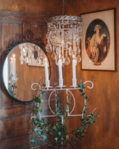 A vintage crystal chandelier and a white iron candelabra standing in front of a round mirror on a warm wooden wall.
