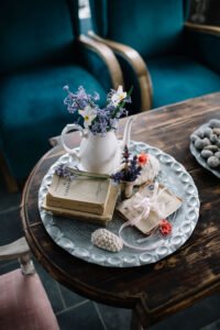 A white vintage teapot with fresh purple lavender on a rustic wooden tray, placed next to old antique books on a dark wooden table.