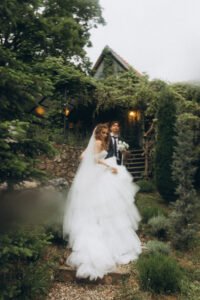 A bride in a flowing tiered gown and a groom holding white flowers looking back from a stone path, surrounded by lush pine trees and a warm, vine-covered cottage at Fairy Oak estate in Fruška Gora.