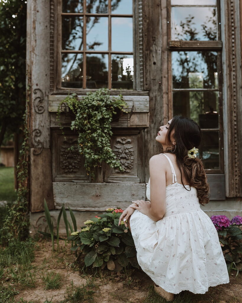 A woman in a white dress kneels in front of the rustic, weathered wooden exterior of the Antik Atelje, surrounded by hydrangeas at Fairy Oak in Vrdnik, Serbia.