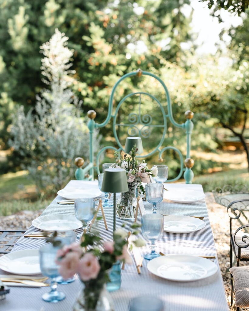 Blue Vintage Garden Wedding Table Setting | Fairy Oak An outdoor wedding table decorated with a blue and white striped tablecloth, blue glass goblets, modern mini green table lamps, and a statement vintage mint-green iron chair at the head of the table.