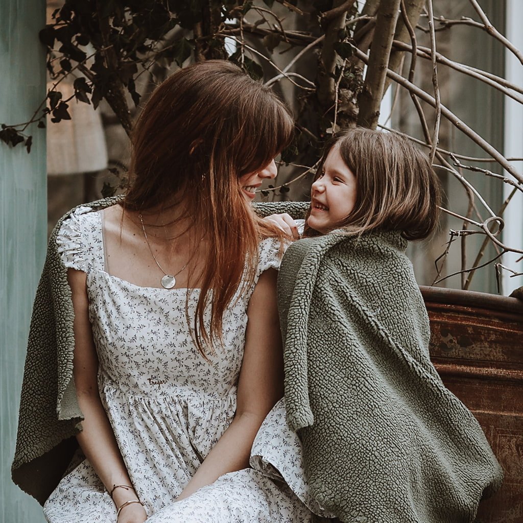 cozy-mother-daughter-portrait-fairy-oak-fruska-gora.jpg A mother and young daughter sit closely together, smiling and laughing warmly at each other. They are wrapped in cozy olive-green fleece blankets, sitting next to a rusted metal structure with bare branches at Fairy Oak in Fruška Gora.