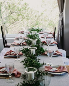 A beautifully set long dining table positioned next to a large bright window, styled with a lush greenery garland runner, white pillar candles in glass cylinders, and mauve napkins at the Fairy Oak estate in Vrdnik.