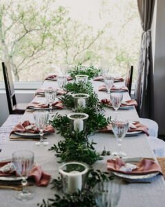 A beautifully set long dining table positioned next to a large bright window, styled with a lush greenery garland runner, white pillar candles in glass cylinders, and mauve napkins at the Fairy Oak estate in Vrdnik.