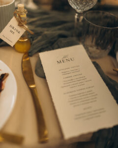 A close-up of an elegant wedding table setting featuring a printed menu, a gold knife resting on a dark green linen napkin, and a small favor bottle with a tag that says "Drink Me".