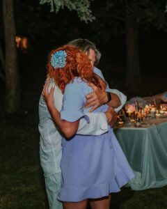 A warm outdoor evening photograph of the Fairy Oak founders sharing a tight hug. A woman with bright curly orange hair wearing a light blue dress embraces a man, with a softly lit event table visible in the background.