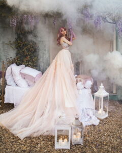 A woman with pink hair wearing a magnificent blush pink Fairy Oak tulle ballgown, standing in a magical, smoke-filled outdoor setting and reaching up to touch hanging purple wisteria. Photographed by Jovana Rikalo.