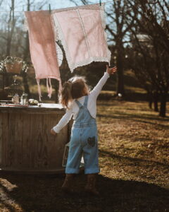 A little girl seen from behind, wearing a white long-sleeve shirt and Fairy Oak denim overalls featuring a floral heart patch on the pocket. She is reaching up towards pink, lace-trimmed linens hanging on an outdoor clothesline in the sunlight. Photographed by Ana Markovic.