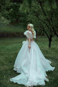 A smiling blonde woman walking through a lush green orchard, looking back over her shoulder. She is wearing a flowing, voluminous white Fairy Oak gown with sheer puffed sleeves and a lace-up corset back. Photographed by Jovana Rikalo.