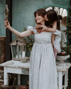 Serbian actress Tamara Dragicevic and a little girl wearing matching white floral Fairy Oak dresses. The girl is playfully hugging Tamara from behind as Tamara smiles into a vintage silver hand mirror. Photographed by Ana Markovic.