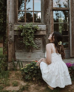 A young woman crouching gracefully in a garden next to blooming pink hydrangeas, looking upwards with her eyes closed. She is wearing a delicate white Fairy Oak sundress with pink floral prints and a smocked back. Photographed by Ana Markovic.