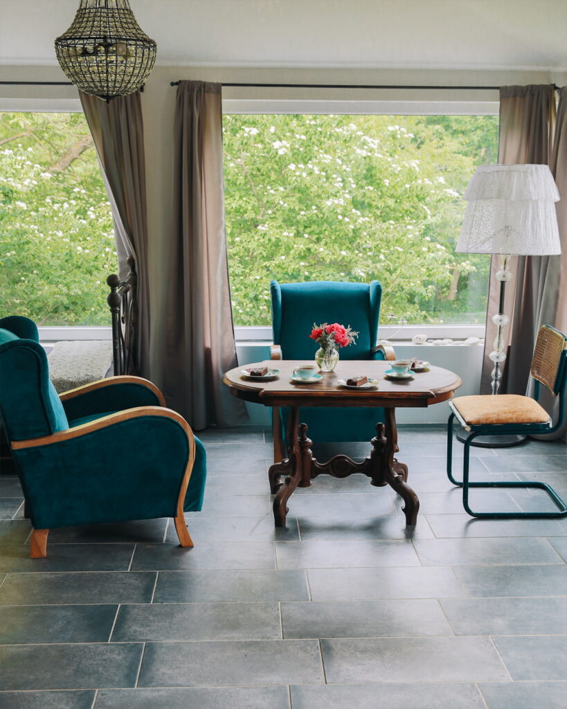 A bright and inviting sunroom interior at Fairy Oak House. A rustic wooden table is set with tea and cake, surrounded by two teal velvet armchairs and a small vintage chair. The room features a crystal chandelier, a floor lamp with a lace shade, and large wall-to-wall windows offering a panoramic view of vibrant green trees outside.
