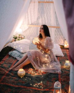 A woman sitting comfortably on a patterned rug inside a cozy sheer tent at night, wearing a lavender Fairy Oak dress with delicate floral embroidery and draped in warm string lights. Photographed by Jovana Rikalo.