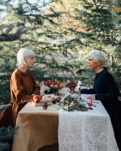 Two women with short blonde hair sitting across from each other at a beautifully styled rustic table in a pine forest. One wears a brown long-sleeved Fairy Oak dress, and the other wears a dark blue Fairy Oak dress. Photographed by Jovana Rikalo.