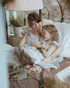 Serbian actress Tamara Dragicevic and a little girl sitting cozily on a bed, smiling and hugging each other affectionately. They are wearing matching white Fairy Oak dresses with delicate floral prints and ruffled sleeves. The candid photo appears to be taken through a window with soft, warm indoor lighting. Photographed by Ana Markovic.