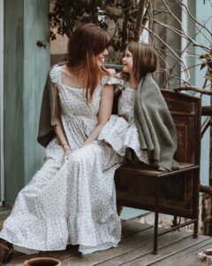 Serbian actress Tamara Dragicevic and a little girl smiling at each other while sitting on a rustic outdoor bench, wearing matching white floral Fairy Oak dresses with a cozy green blanket draped over their shoulders. Photographed by Ana Markovic.