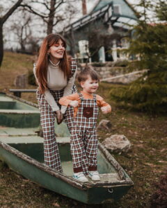 Serbian actress Tamara Dragicevic wearing plaid wide-leg pants and a little boy in matching Fairy Oak plaid overalls standing playfully on a green wooden boat outdoors in a garden setting. Photographed by Ana Markovic.