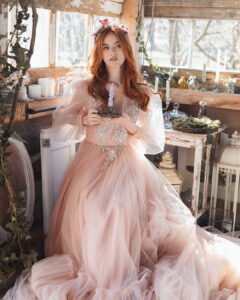 A red-haired woman wearing a voluminous peach tulle Fairy Oak fantasy gown with sheer puffed sleeves and silver appliqué, sitting in a rustic sunlit room holding a lit purple taper candle. Photographed by Jovana Rikalo.