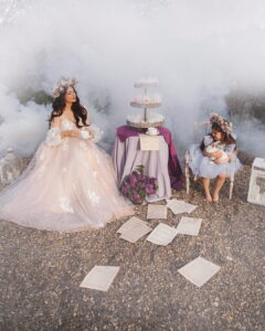 Serbian fashion blogger Ena Luna wearing a voluminous pale pink Fairy Oak tulle gown with white floral appliques and a flower crown, holding a teacup at a magical, smoke-filled outdoor tea party. Next to her, a little girl in a blue tulle dress sits holding a white rabbit, with vintage sheet music scattered on the ground. Photographed by Jovana Rikalo.
