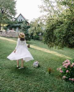 A woman seen from behind, wearing a straw boater hat and a white midi-length Fairy Oak dress, twirling on a lush green lawn. The idyllic garden setting features a quaint blue cottage with a stone wall, large trees, and pink hydrangeas in the foreground. Photographed by Jovana Rikalo.