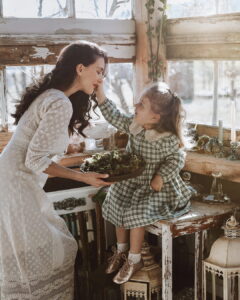A woman wearing a long-sleeved white lace Fairy Oak dress holding a tray of moss, smiling warmly as a little girl in a green plaid dress reaches out to touch her face inside a sunlit rustic greenhouse. Photographed by Ana Markovic.