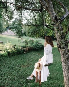 A woman with wavy brown hair sitting on a wooden tree swing in a lush green garden, seen from behind. She is wearing a long-sleeved white Fairy Oak linen dress with a back tie and holding a straw hat with a brown ribbon. Photographed by Jovana Rikalo.