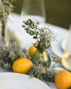 A close-up of an outdoor wedding table decoration featuring fresh lemons, textured dusty miller foliage, and a small green vintage vase holding sprigs of eucalyptus.