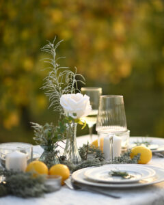 A garden wedding table setting close-up showing fresh whole lemons, sprigs of greenery, a white rose in a small fluted glass vase, and ribbed drinking glassware.