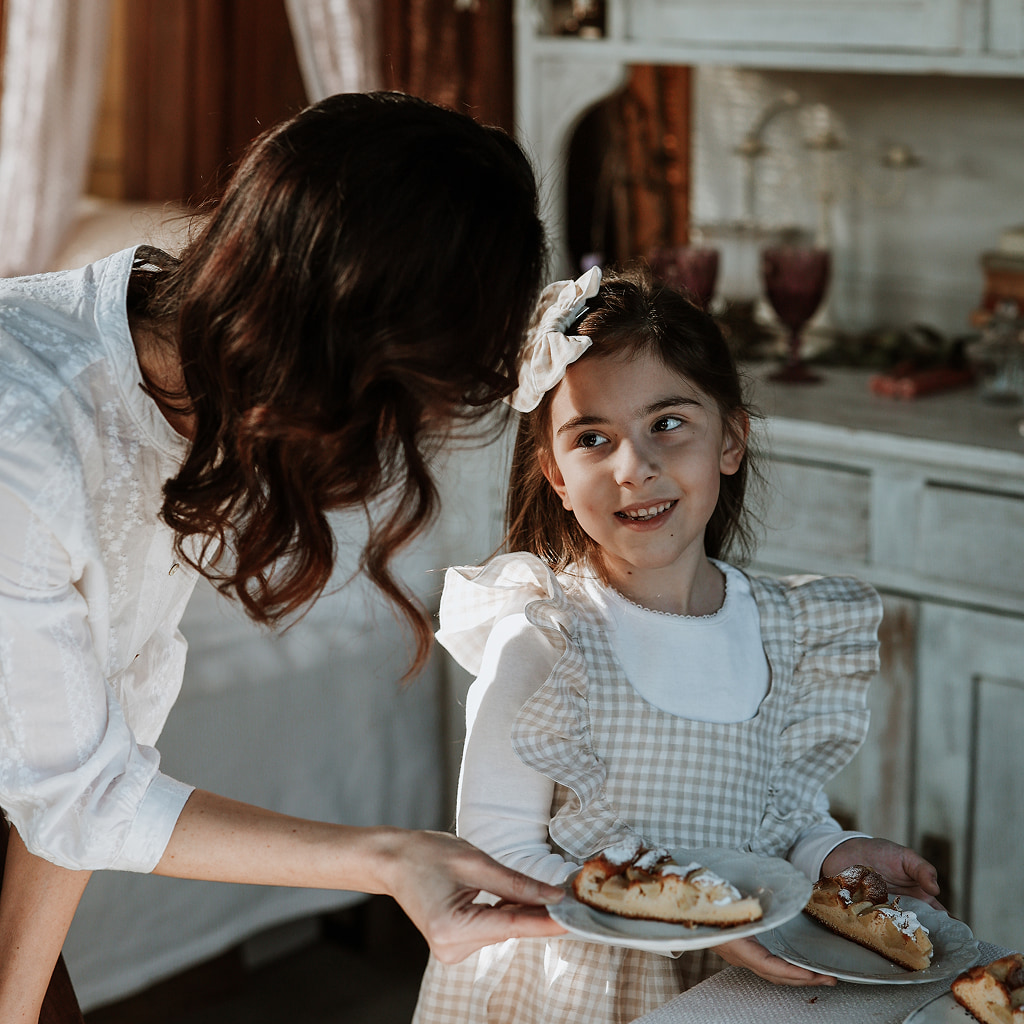 A mother serving a slice of freshly baked pie to her smiling young daughter in a rustic, natural-light kitchen.