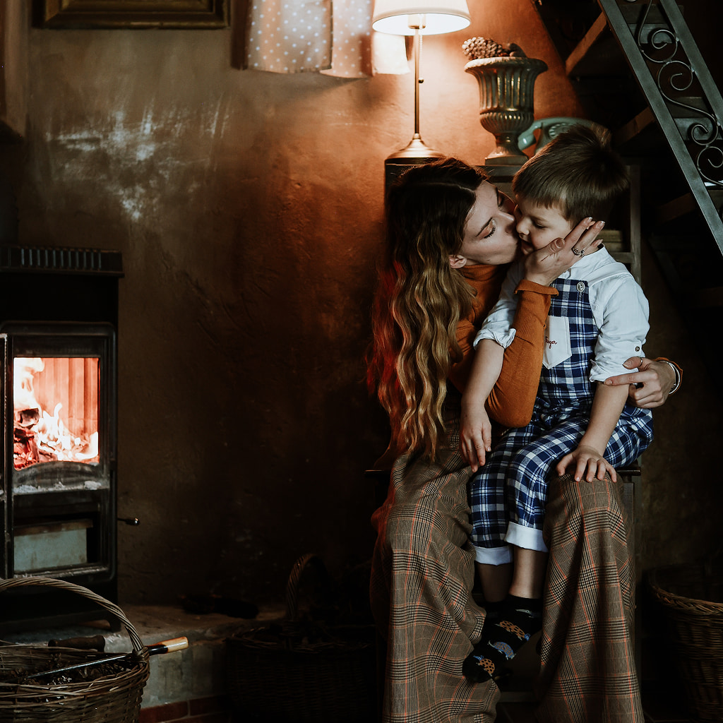 A mother affectionately kissing her young son on the cheek while sitting next to a warm, glowing wood-burning stove in a cozy, rustic room.