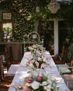 A long banquet table set for a wedding reception on a covered outdoor patio, decorated with pink and white floral centerpieces beneath a rustic crystal chandelier at Fairy Oak, Serbia.