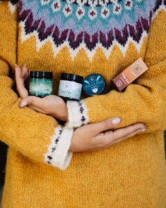 A close-up of a person wearing a cozy, patterned yellow knit sweater holding a collection of Talga Cosmetics products, including face creams, beard balm, and lip balm against their chest.