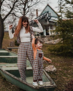 Actress Tamara Dragičević and a little boy playfully model matching Tregertim plaid overalls next to a rustic green boat. The beautiful, distinctive exterior of the Fairy Oak house is visible in the background.