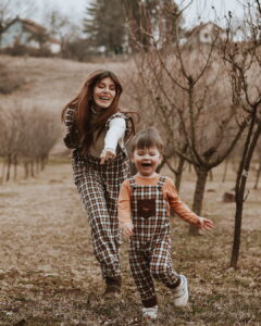 Actress Tamara Dragičević and a young boy run joyfully toward the camera through a dormant orchard, both wearing matching Treger brand plaid overalls during a lifestyle fashion shoot at Fairy Oak.