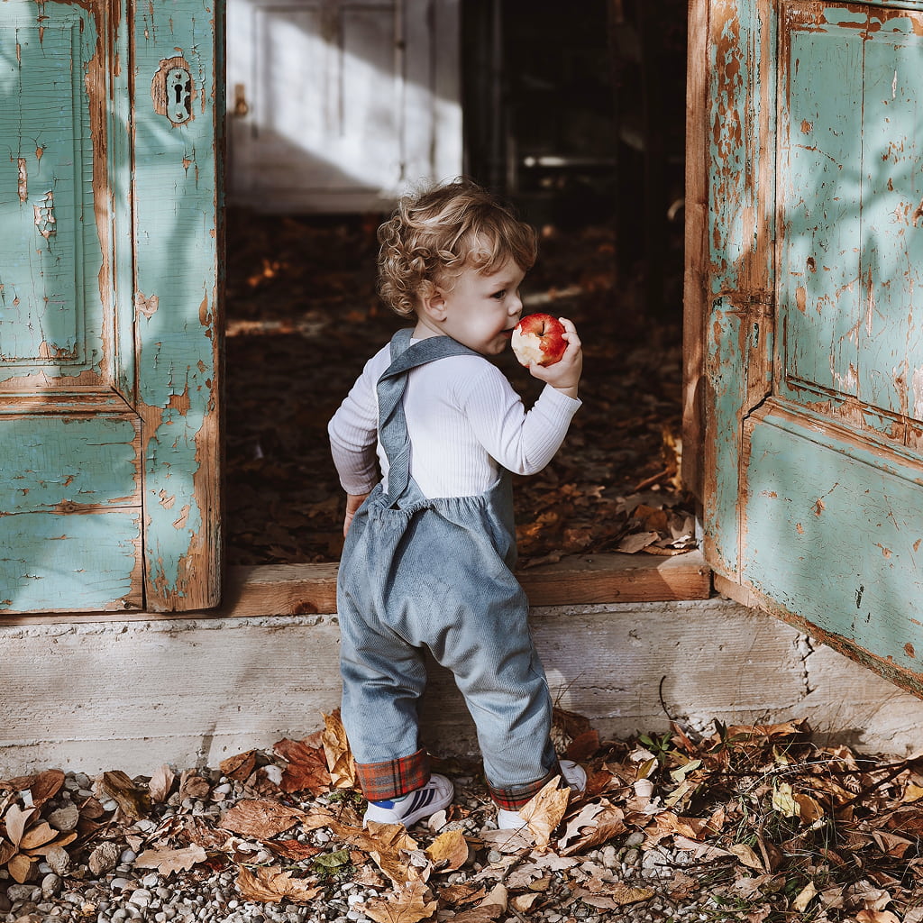 A curly-haired toddler wearing blue corduroy overalls stands in the doorway of weathered teal wooden doors, eating a red apple. The ground is covered in crisp autumn leaves during a fall photo session at Fairy Oak in Fruška Gora.