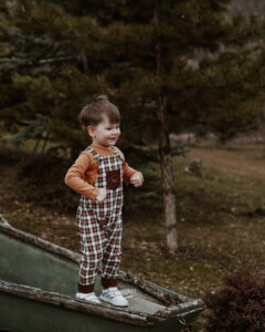 A smiling little boy wearing Tregertim brand brown and white plaid overalls stands playfully on a rustic green wooden boat during an autumn children's fashion photo shoot at Fairy Oak.