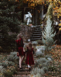 Two young girls wearing matching red velvet Treger dresses with ruffle details walk hand-in-hand up outdoor stone steps surrounded by autumn foliage and greenery at the Fairy Oak estate.