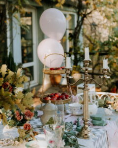 An outdoor event table styled with vintage charm, featuring a brass candelabra, a tiered brass tray of peaches, pale pink balloons, and floral arrangements at Fairy Oak in Vrdnik.