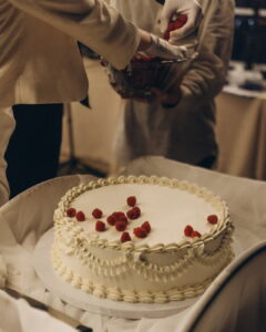 A person wearing white gloves carefully places fresh red raspberries onto a classic white frosted wedding cake decorated with elegant vintage-style piping.