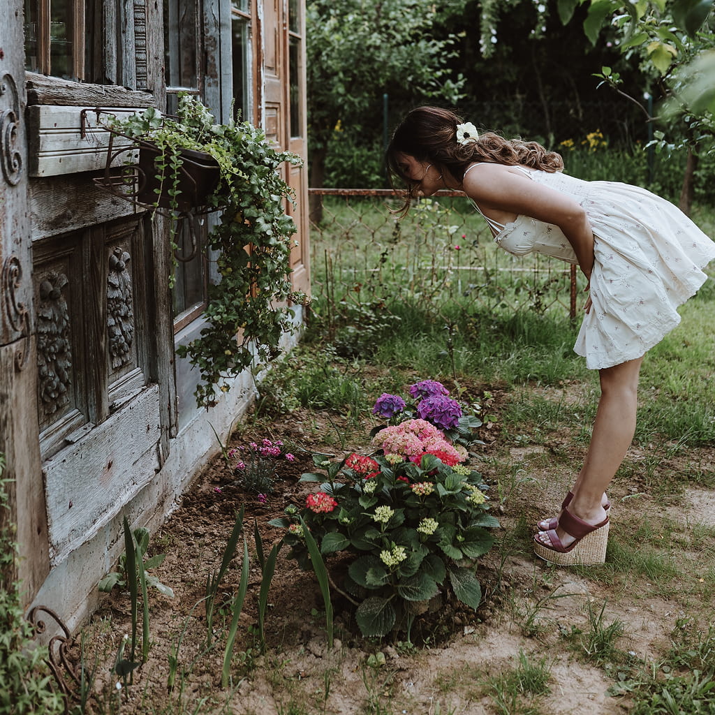 A woman wearing a short white sundress and maroon platform sandals leans over to admire small pink and purple hydrangeas planted in the dirt. She is standing outdoors in the vintage garden at Fairy Oak in Fruška Gora, next to an ornate wooden building.